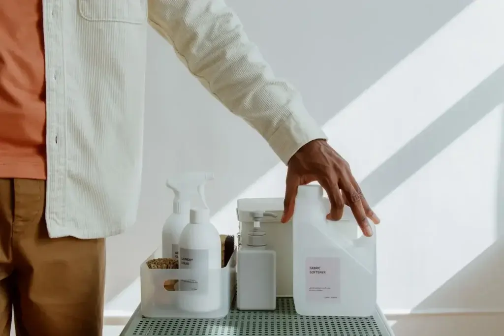 Nahaufnahme eines Mannes, der einen Weichspüler in der Hand hält, umgeben von Reinigungsmitteln in einer minimalistischen Umgebung.