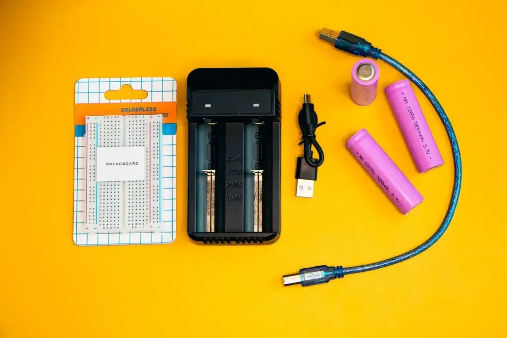 Fotografía de productos electrónicos en plano, entre los que se incluyen una placa de pruebas y un cargador de baterías, sobre un fondo de color amarillo intenso.