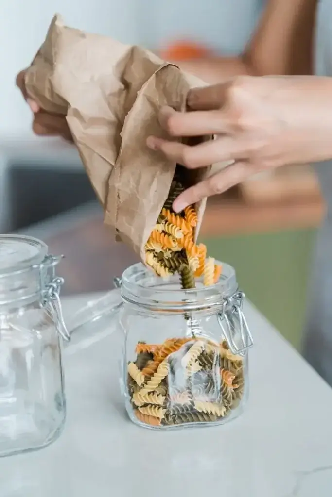 Close-up of a person transferring tri-color pasta from a paper bag into a glass jar, indoors.